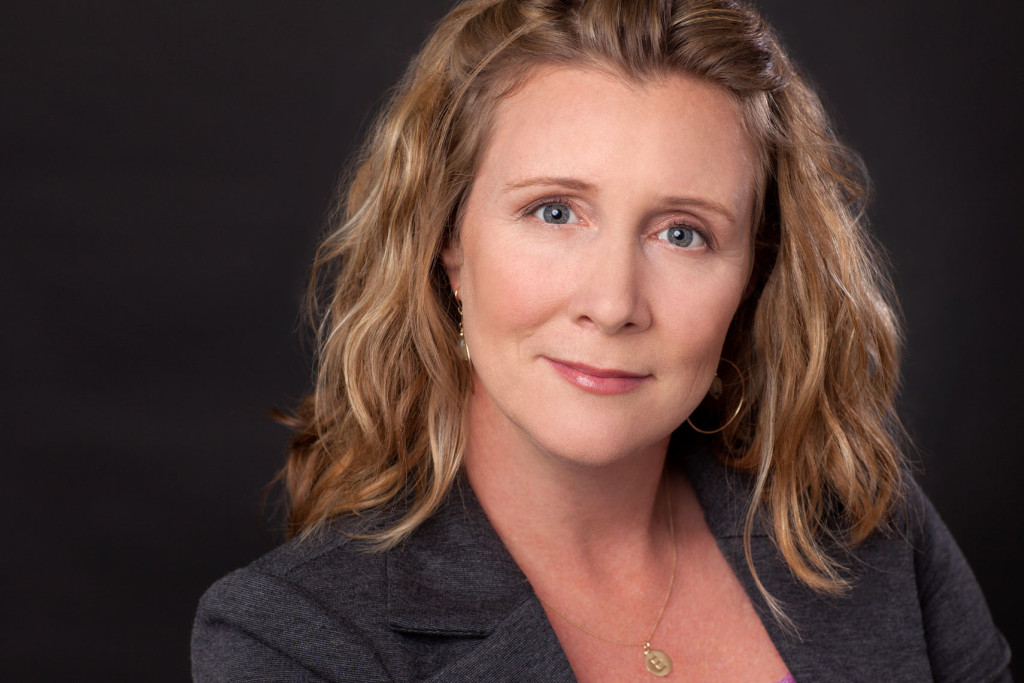 Go-To Expression headshot of blonde woman smizing. She wears a grey blazer and gold necklace with an E on it. Black Background.