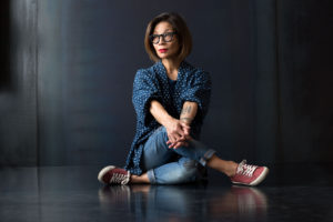 Portrait of Woman with glasses and arm tattoo sitting cross legged in front of a metal background.