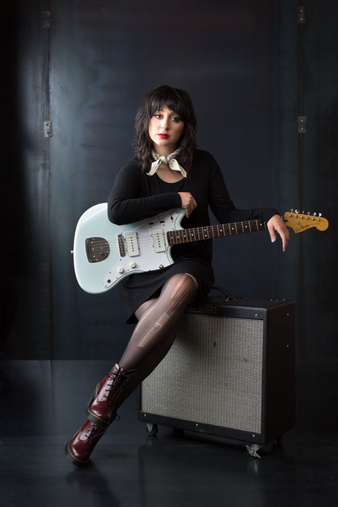 Woman Musician sits on an amp with her guitar